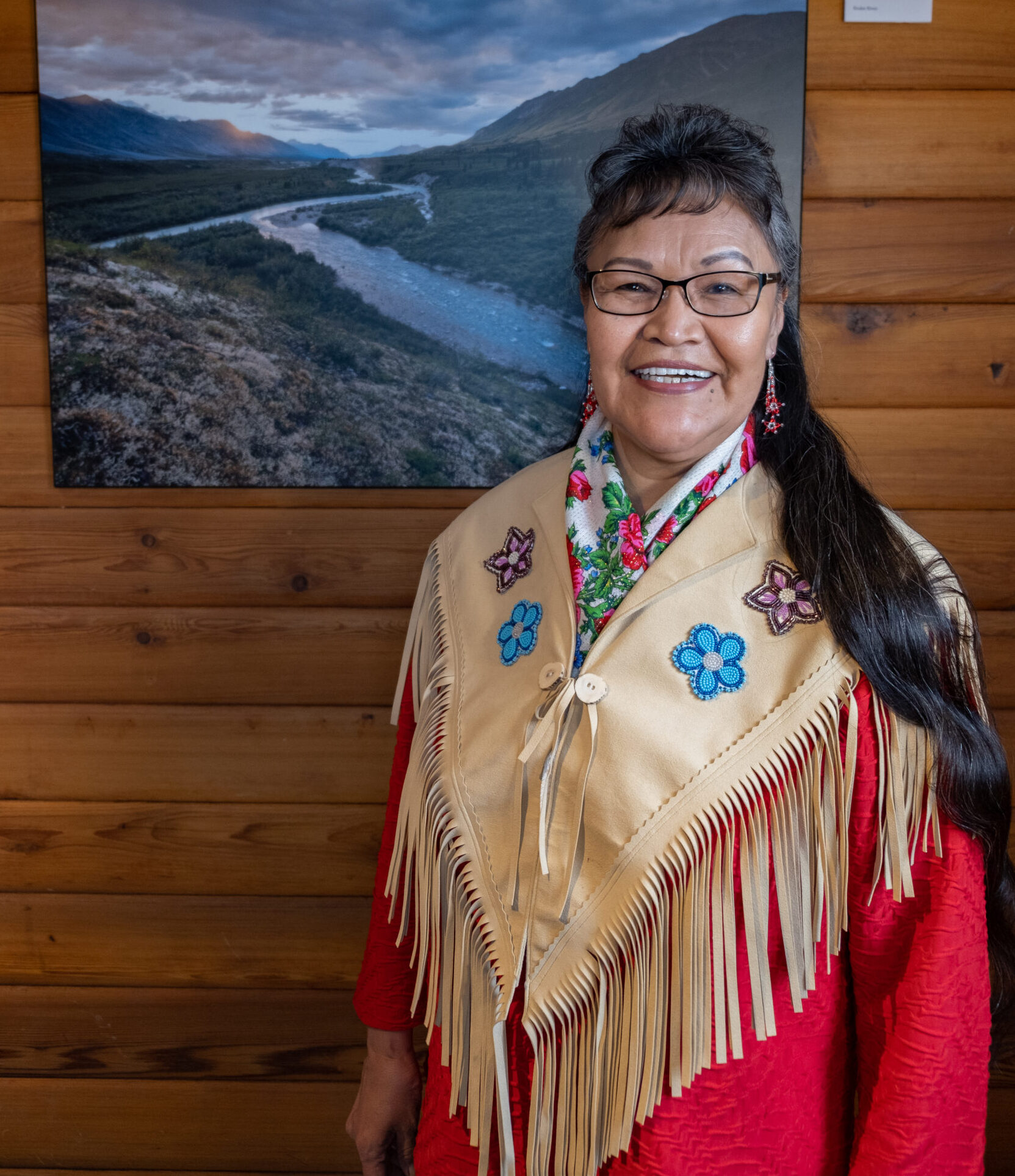 Smiling Indigenous woman in traditional beaded leather shawl stands beside a photo of the Yukon landscape