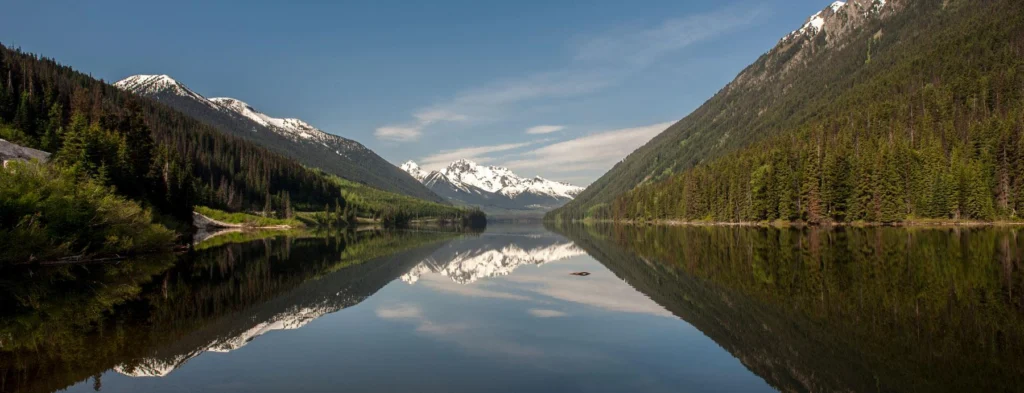 Wide-angle photography of a forested mountain valley against a blue sky with a lake reflecting the scene below.
