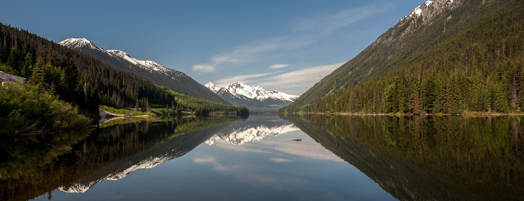 Wide-angle photography of a forested mountain valley against a blue sky with a lake reflecting the scene below.